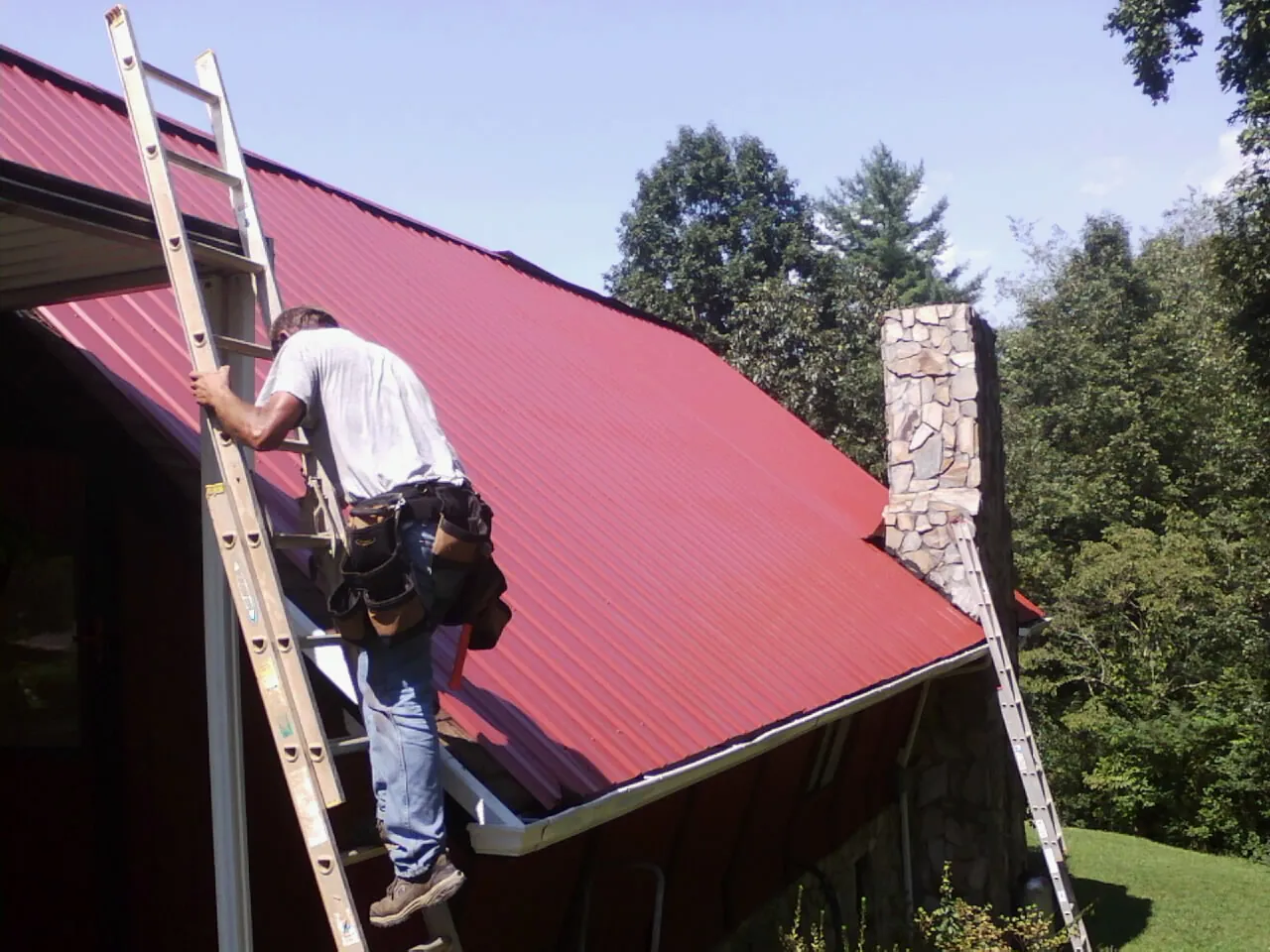Roofer on a ladder beside a red metal roof and stone chimney — Soffit & Fascia Repair by Roofing Brothers in Buffalo Prairie